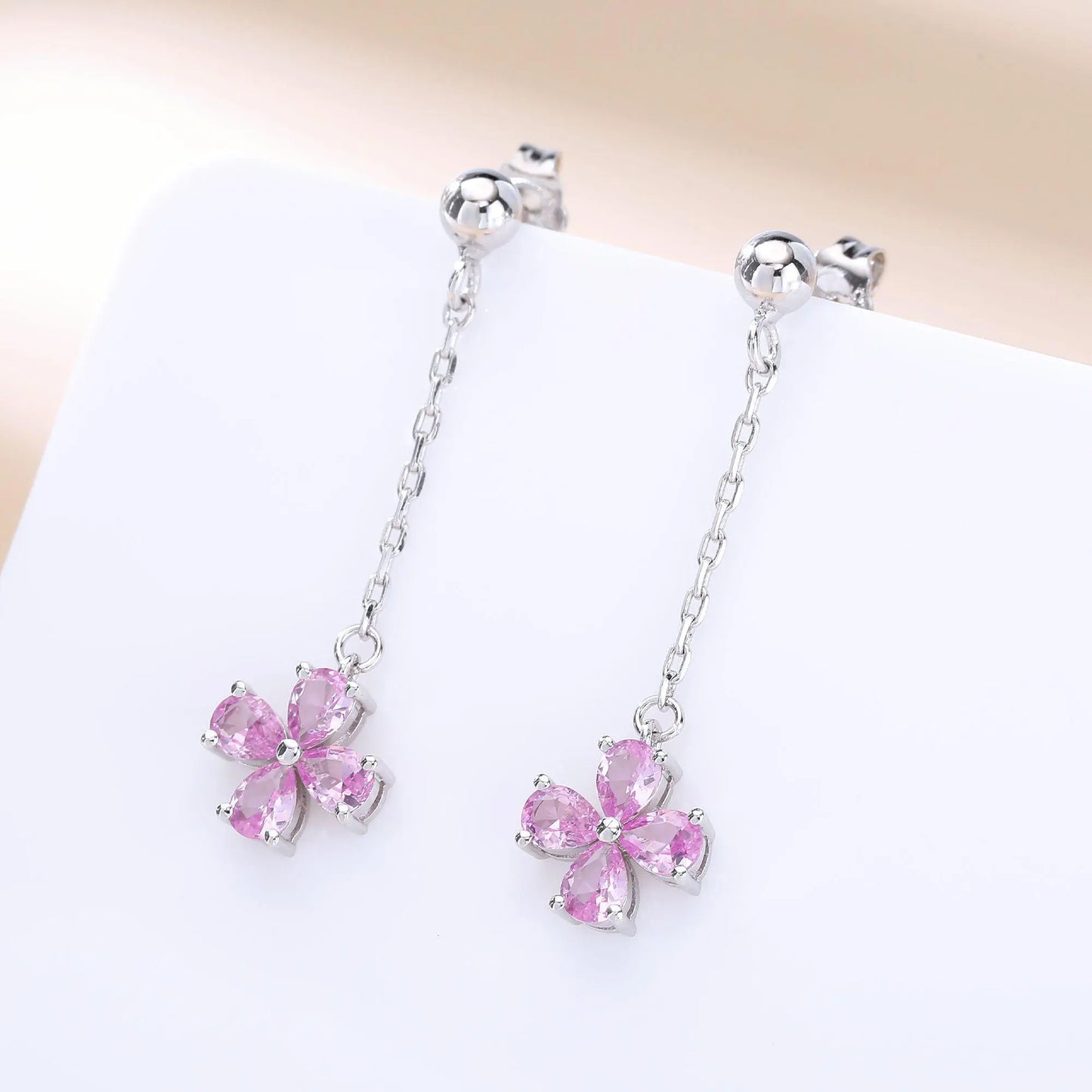 A pair of sterling silver drop stud earrings featuring a shimmering four-leaf clover charm made of four pear-cut pink sapphire gemstones, suspended from a delicate silver chain.