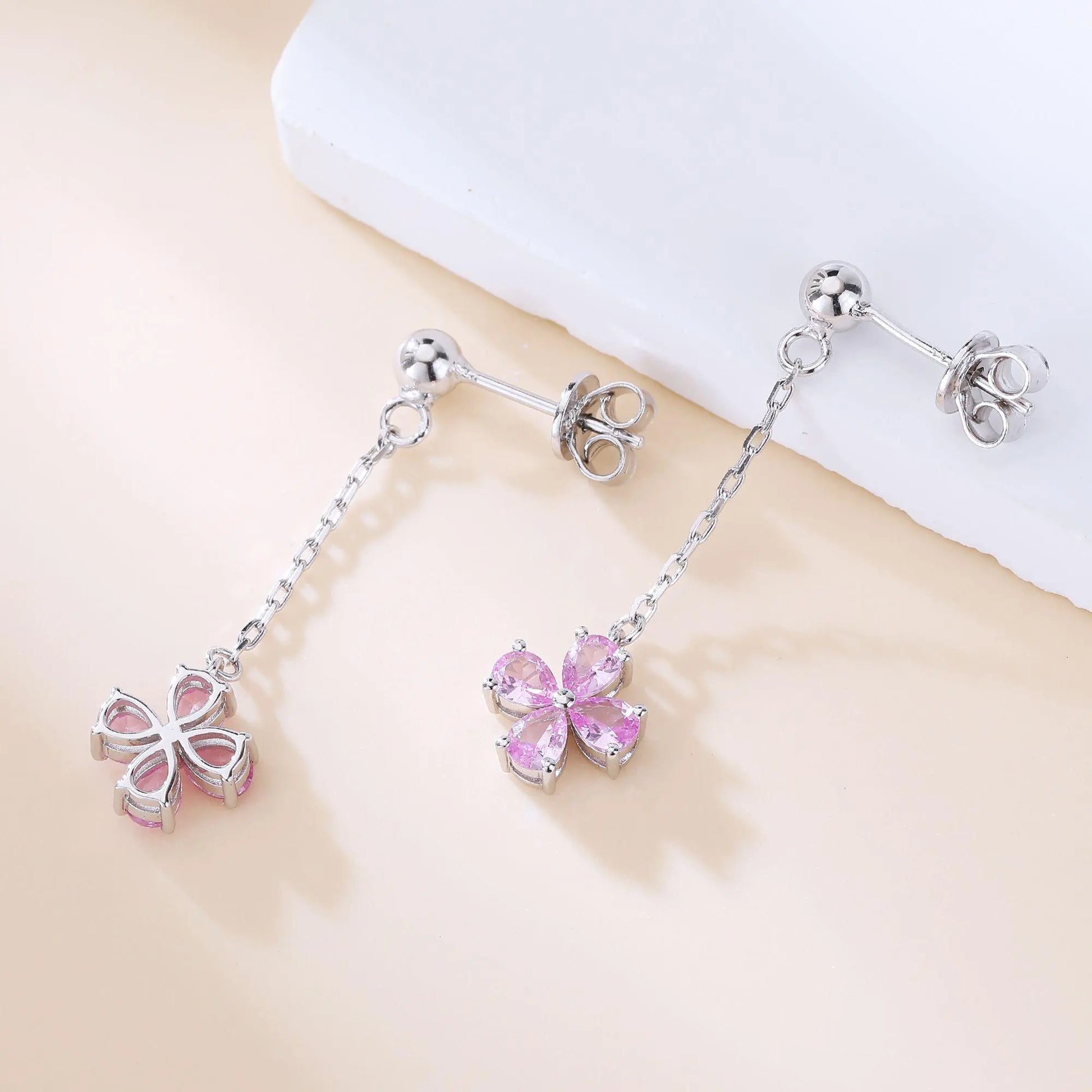 A pair of sterling silver drop stud earrings featuring a shimmering four-leaf clover charm made of four pear-cut pink sapphire gemstones, suspended from a delicate silver chain.