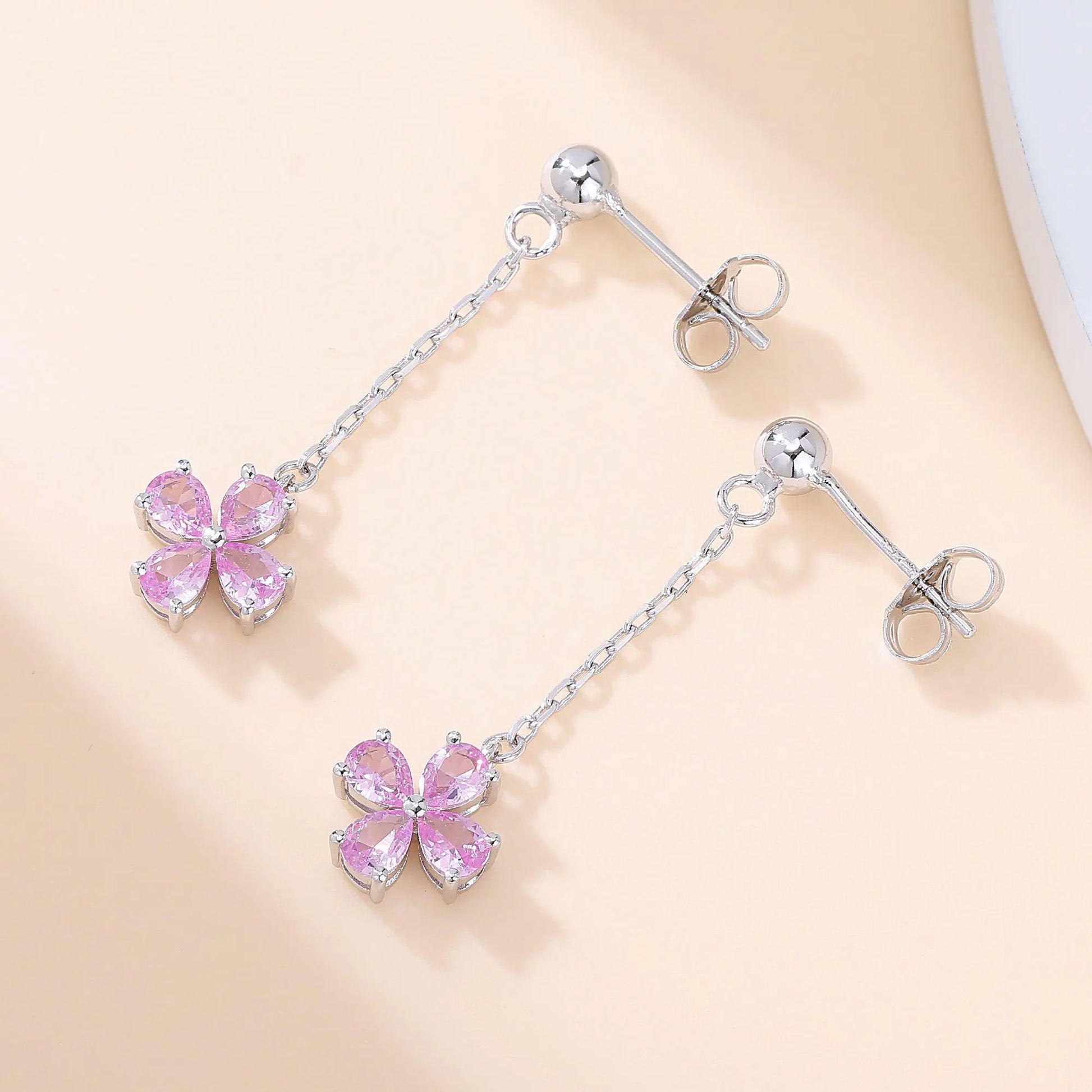 A pair of sterling silver drop stud earrings featuring a shimmering four-leaf clover charm made of four pear-cut pink sapphire gemstones, suspended from a delicate silver chain.