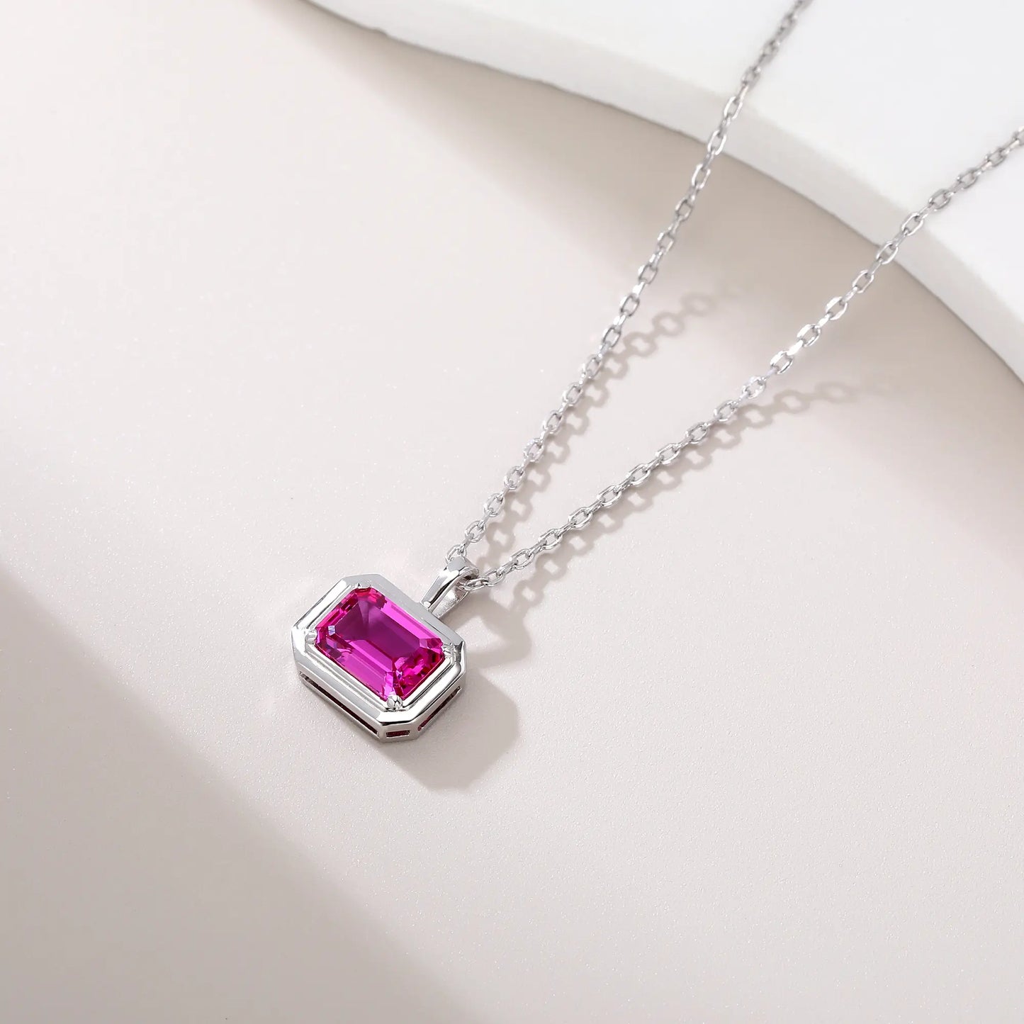 A horizontal emerald cut pink sapphire pendant necklace with a sleek sterling silver bezel setting, showing vibrant pink facets and a reflective white background.