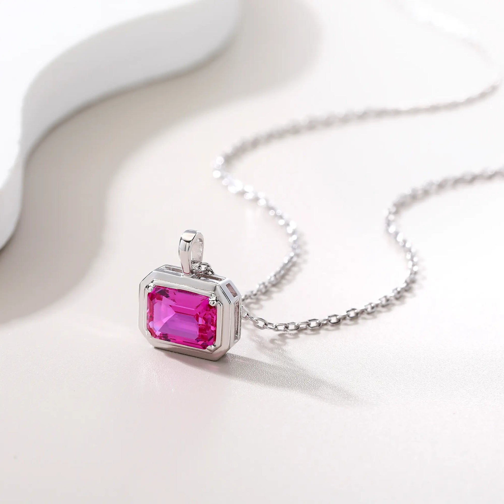A horizontal emerald cut pink sapphire pendant necklace with a sleek sterling silver bezel setting, showing vibrant pink facets and a reflective white background.