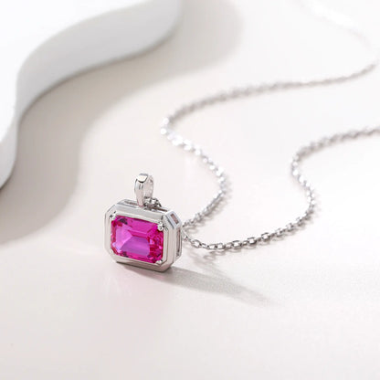 A horizontal emerald cut pink sapphire pendant necklace with a sleek sterling silver bezel setting, showing vibrant pink facets and a reflective white background.
