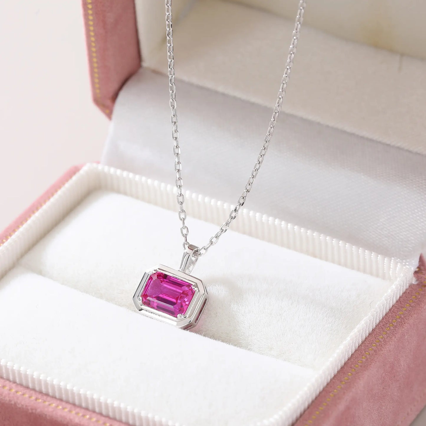 A horizontal emerald cut pink sapphire pendant necklace with a sleek sterling silver bezel setting, showing vibrant pink facets and a reflective white background.