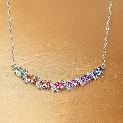 A curved rainbow pendant necklace featuring seven oval cut multi-color sapphires separated by diamond-encrusted silver swirl links, set against a crisp white background.