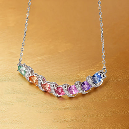 A curved rainbow pendant necklace featuring seven oval cut multi-color sapphires separated by diamond-encrusted silver swirl links, set against a crisp white background.
