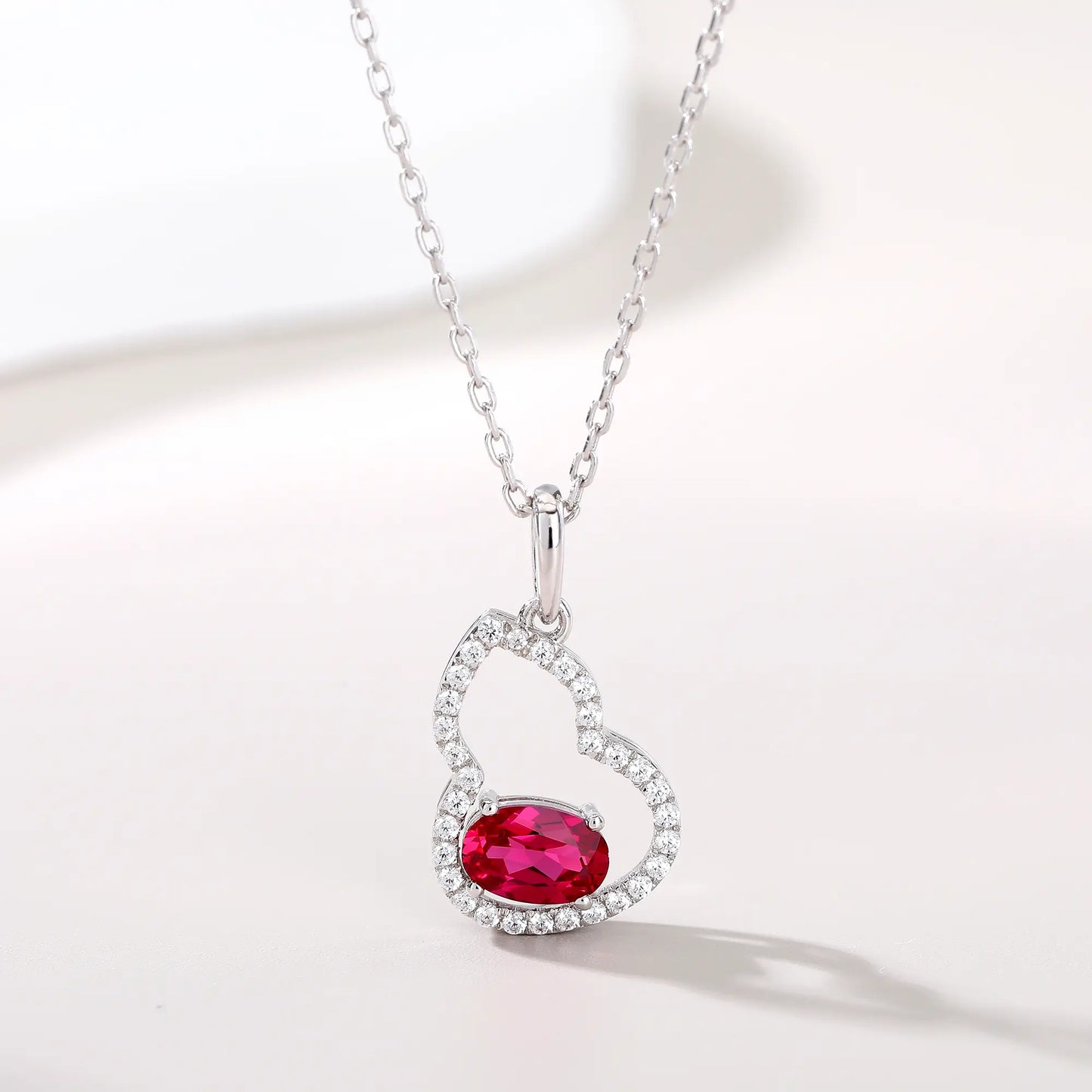An openwork gourd pendant necklace paved with sparkling cz stones, featuring a horizontally set oval cut ruby at the base, resting on a white reflective surface.