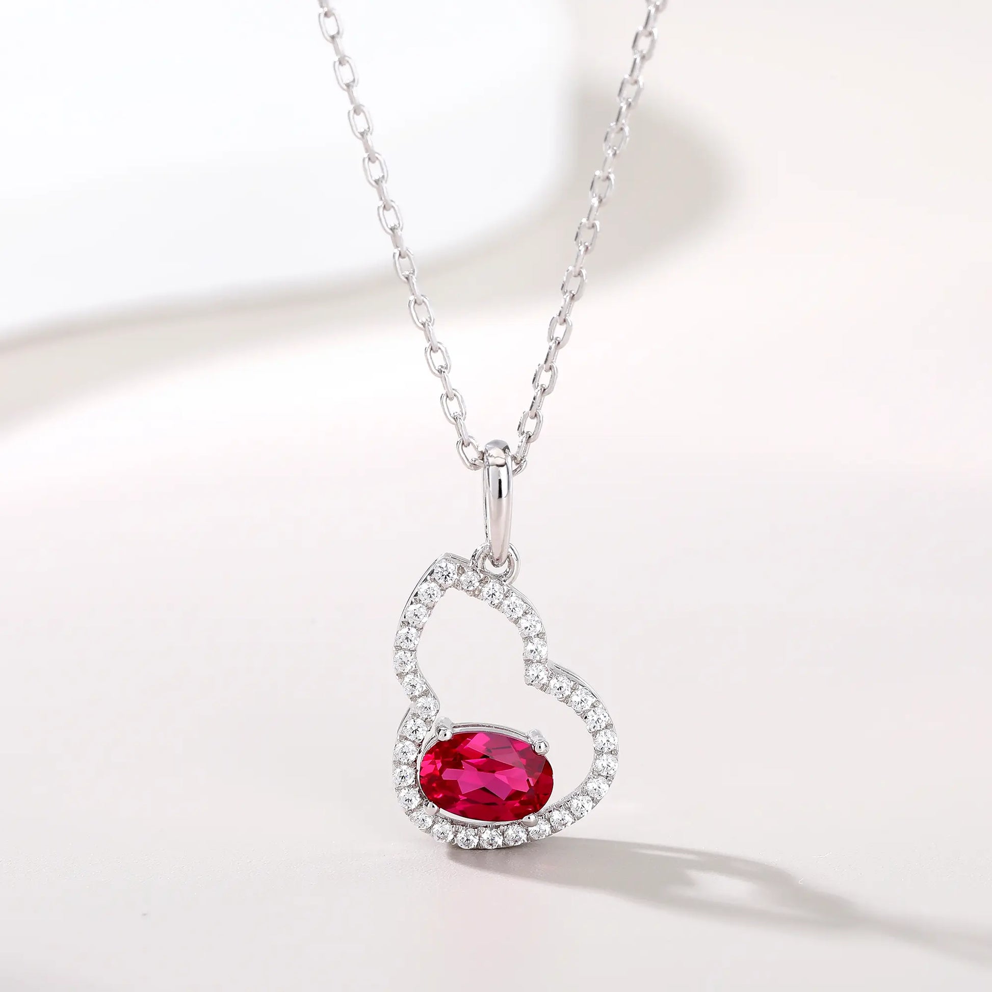 An openwork gourd pendant necklace paved with sparkling cz stones, featuring a horizontally set oval cut ruby at the base, resting on a white reflective surface.