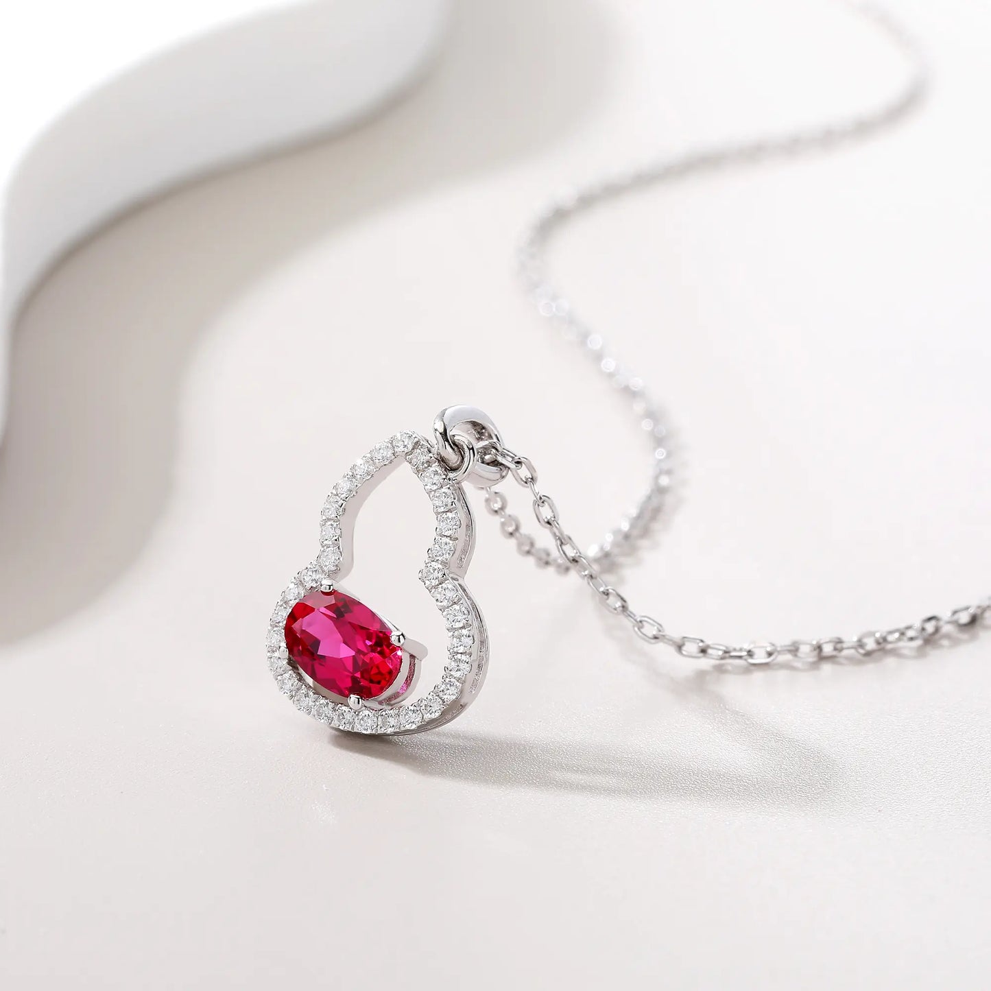 An openwork gourd pendant necklace paved with sparkling cz stones, featuring a horizontally set oval cut ruby at the base, resting on a white reflective surface.
