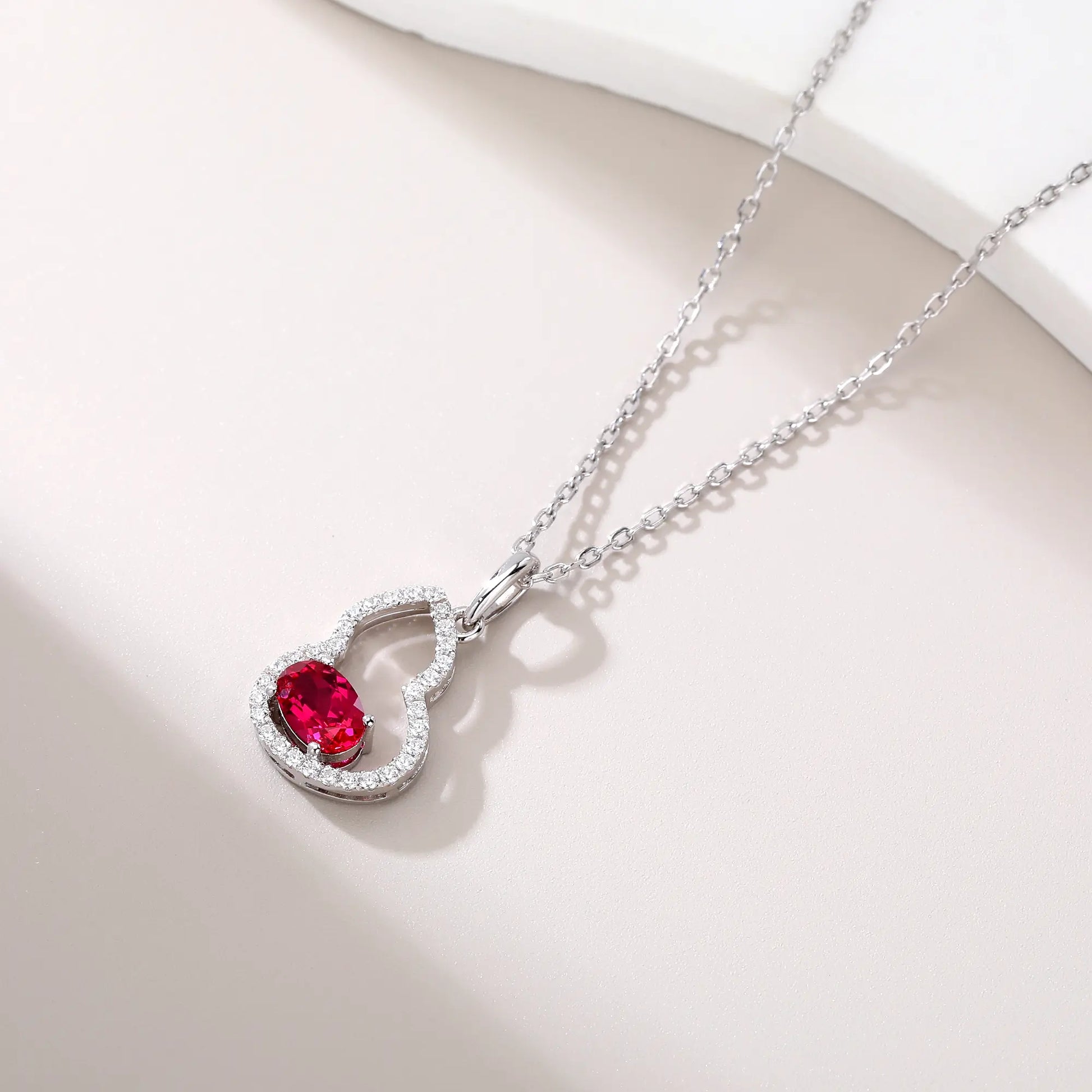 An openwork gourd pendant necklace paved with sparkling cz stones, featuring a horizontally set oval cut ruby at the base, resting on a white reflective surface.