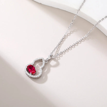 An openwork gourd pendant necklace paved with sparkling cz stones, featuring a horizontally set oval cut ruby at the base, resting on a white reflective surface.