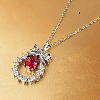 A delicate sterling silver pendant necklace featuring an elegant metal bow tied above a sparkling halo wreath, centering a vibrant pear-cut red ruby teardrop that catches the light.