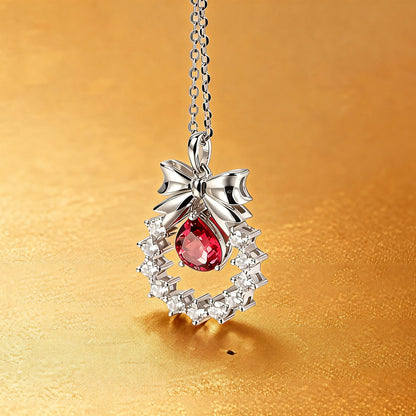 A delicate sterling silver pendant necklace featuring an elegant metal bow tied above a sparkling halo wreath, centering a vibrant pear-cut red ruby teardrop that catches the light.