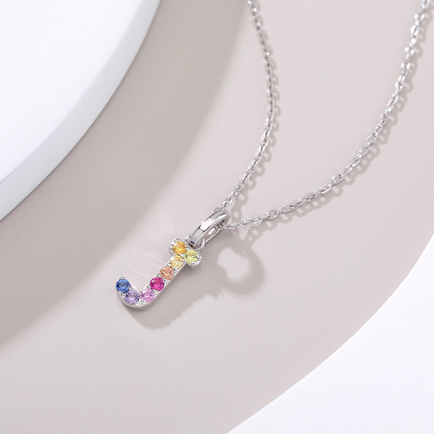 A graceful sterling silver cable chain necklace presenting a capital letter J charm set with a fluid rainbow arc of multi-color lab sapphires.