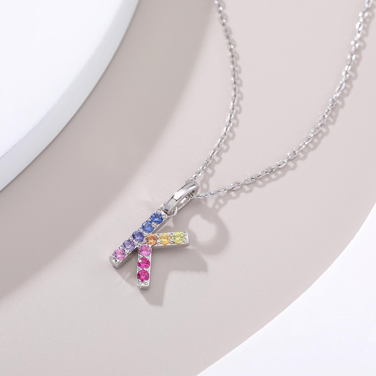 A solid sterling silver necklace displaying a sharp letter K pendant prong-set with a striking multi-color rainbow gradient of brilliant lab sapphires.