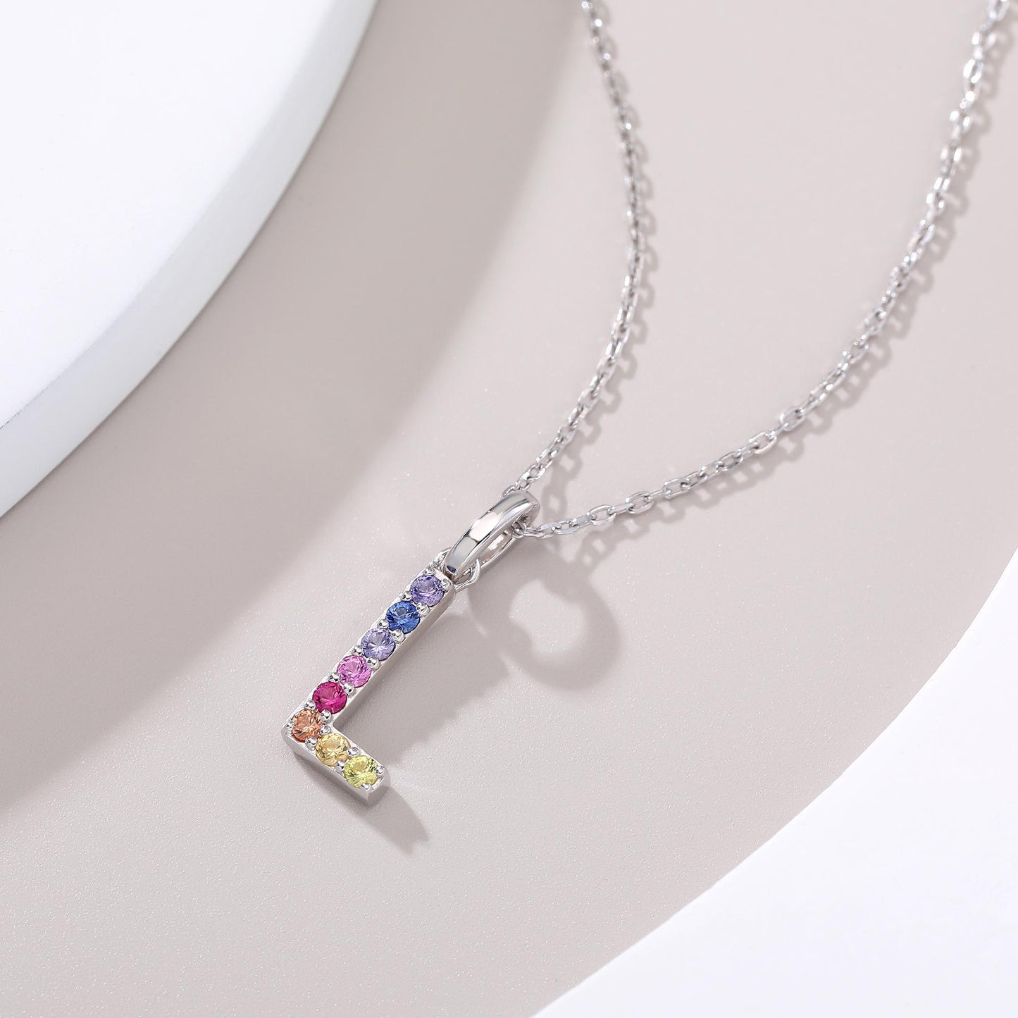 A minimal sterling silver link chain necklace presenting a capital letter L pendant handset with a vibrant vertical and horizontal rainbow layout of multi-color lab sapphires.
