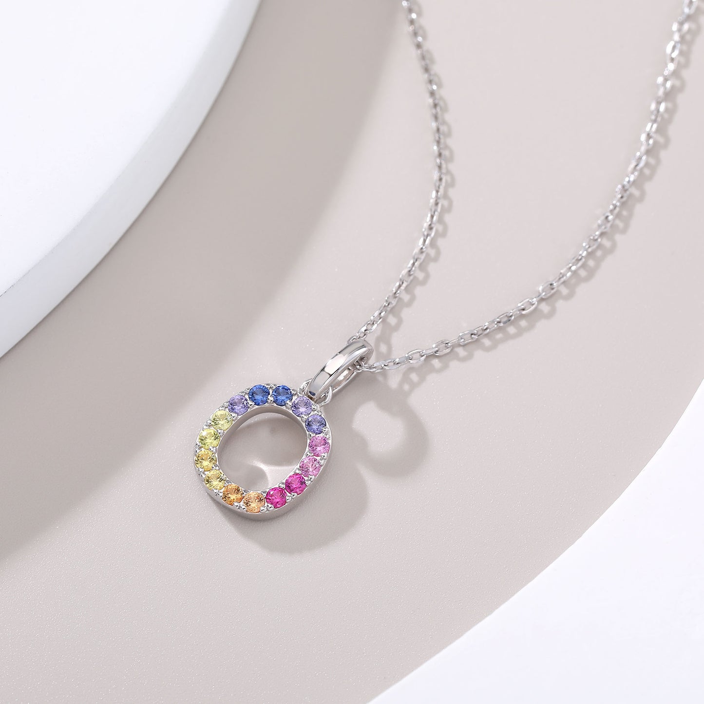 A delicate sterling silver cable chain necklace displaying a circular capital letter O pendant beautifully encrusted with a continuous rainbow gradient of multi-color lab sapphires.