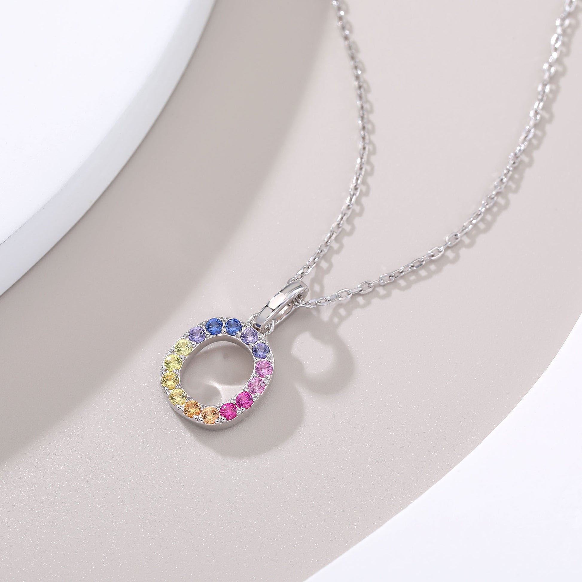 A delicate sterling silver cable chain necklace displaying a circular capital letter O pendant beautifully encrusted with a continuous rainbow gradient of multi-color lab sapphires.