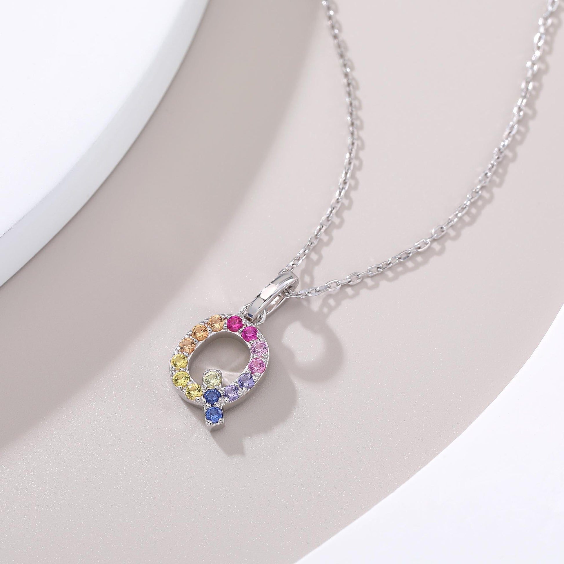A solid sterling silver necklace displaying an uppercase letter Q monogram pendant, highlighted by a playful tail and handset with a continuous rainbow gradient of round lab sapphires.