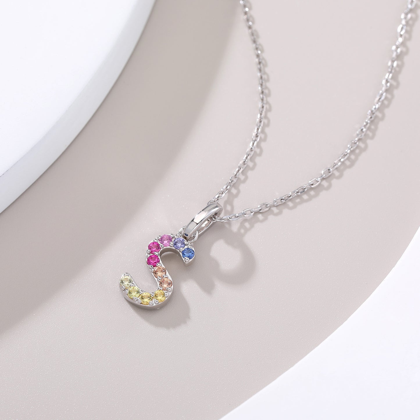 A delicate sterling silver initial necklace showcasing a fluid, serpentine letter S pendant precision-set with a continuous rainbow wave of vibrant round lab sapphires.