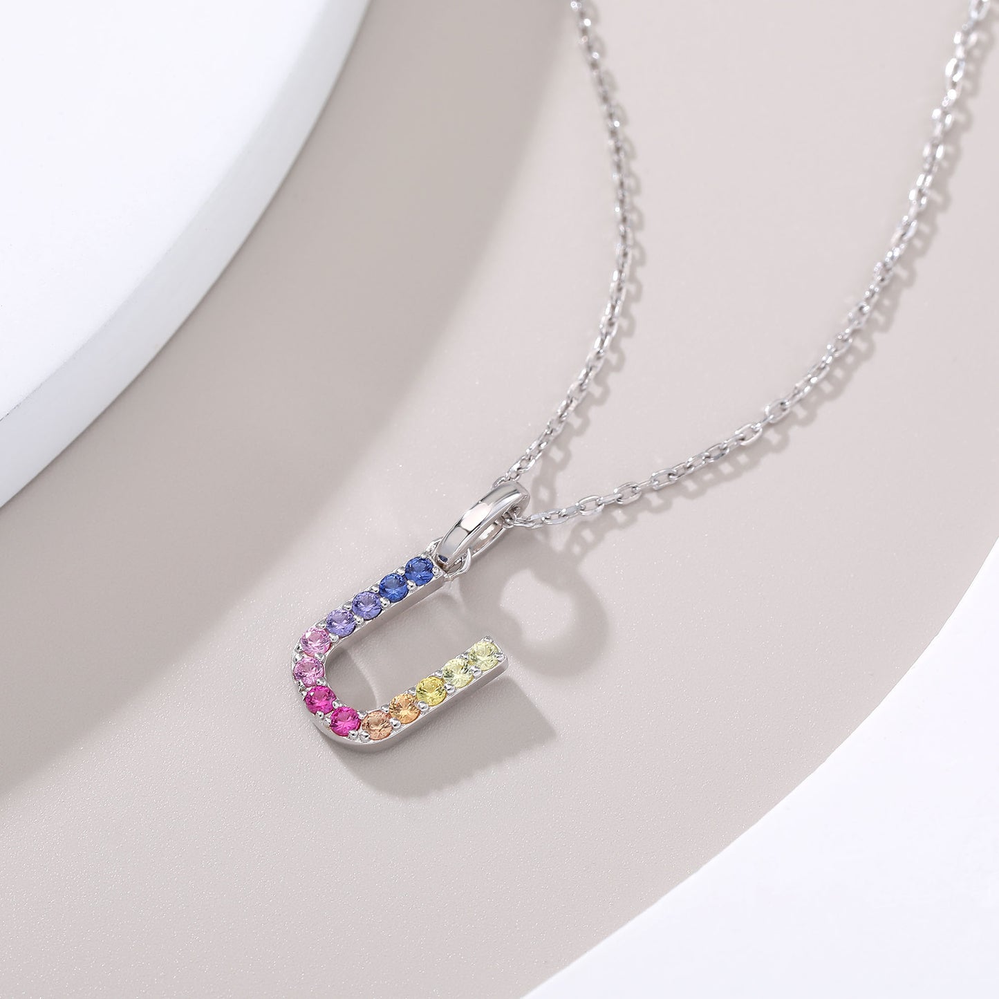 A delicate sterling silver chain necklace suspending an uppercase letter U charm, micro-pave set with a continuous u-shaped rainbow gradient of multi-color lab sapphires.