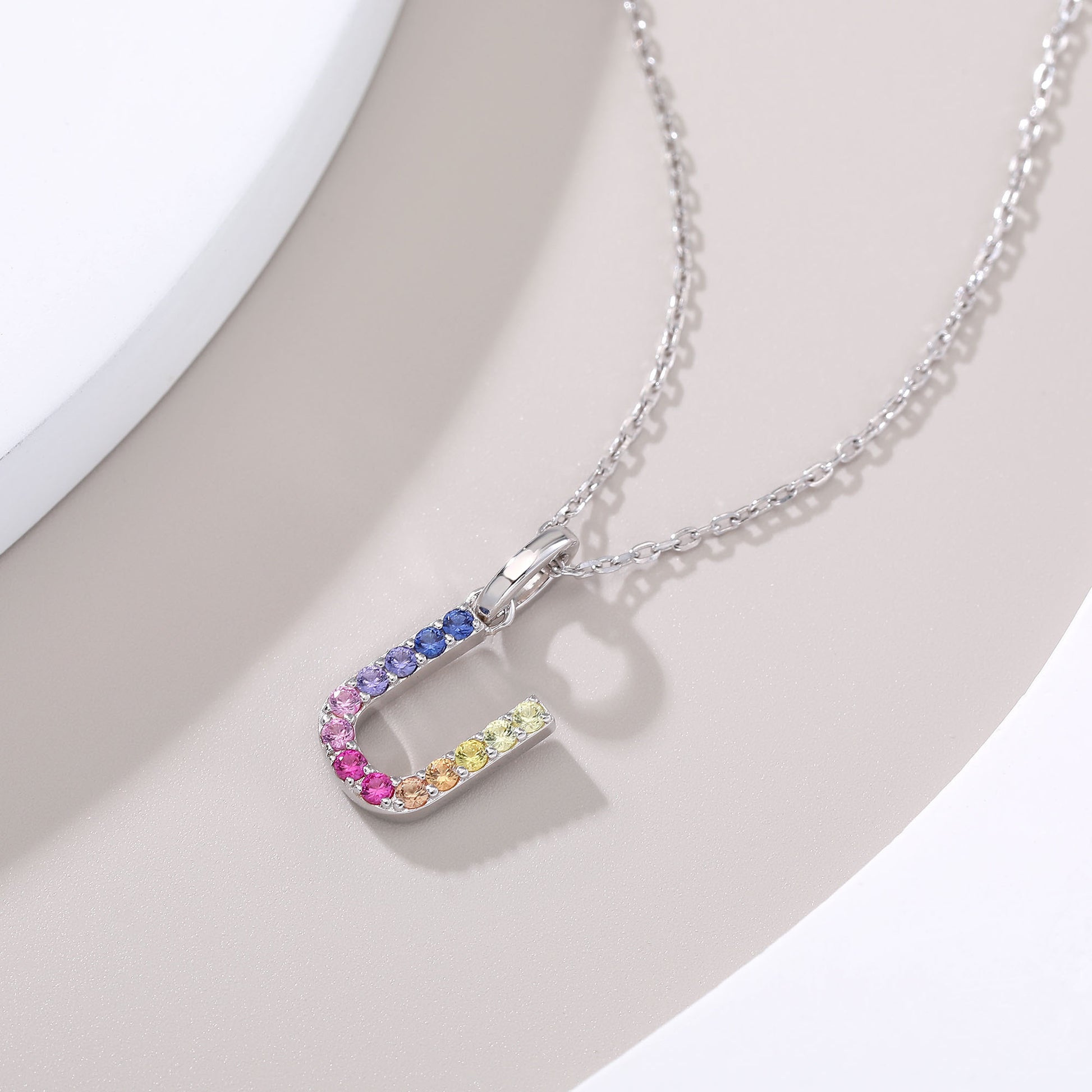 A delicate sterling silver chain necklace suspending an uppercase letter U charm, micro-pave set with a continuous u-shaped rainbow gradient of multi-color lab sapphires.