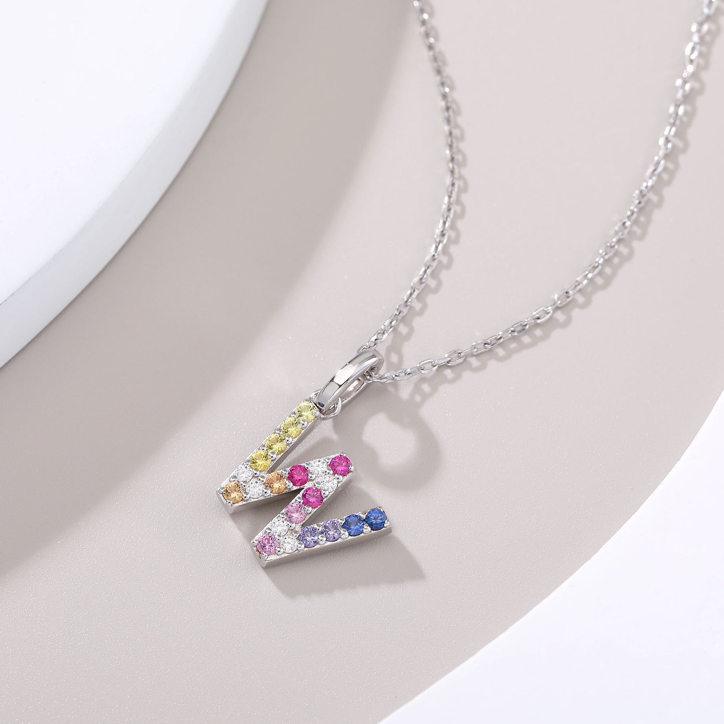 A delicate sterling silver cable chain necklace supporting an uppercase letter W pendant, micro-pave set with a continuous zigzag rainbow gradient of multi-color lab sapphires.